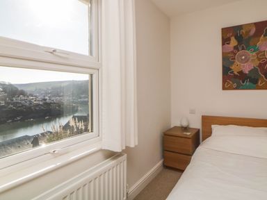 A bedroom with a bed next to a wooden nightstand and a window showing a river view at Estuary View in Newton Ferrers