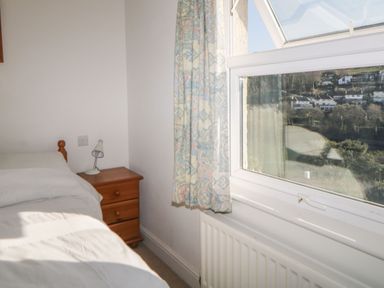 A bedroom corner with a bed a wooden nightstand with a lamp and an open window showing houses outside at Estuary View in Newton Ferrers