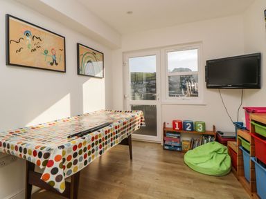 A room with a table covered by a polka dot tablecloth two framed drawings on the wall a wall-mounted television shelves with numbered and colored bins a green bean bag and a door and window showing outside at Estuary View in Newton Ferrers
