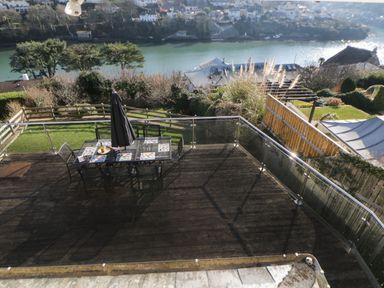 A wooden deck with a glass table and chairs next to a green lawn with trees and a river in the background at Estuary View in Newton Ferrers