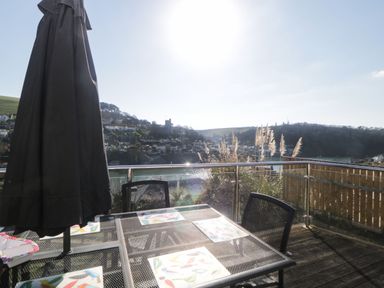 A balcony with a glass railing a metal table with chairs and a closed umbrella overlooking a hillside town and water at Estuary View in Newton Ferrers