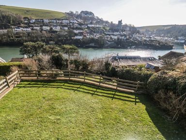 A grassy backyard with a wooden fence overlooking a river and a hillside with houses at Estuary View in Newton Ferrers