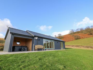 A modern single-story house with wooden bench and hot tub on grassy hill at Dol Blodau in Newbridge-On-Wye