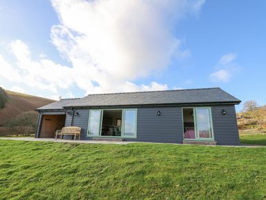 A single story house with sliding glass doors and a wooden bench on a grassy lawn at Dol Blodau in Newbridge-On-Wye