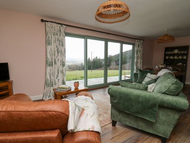 A living room with brown and green sofas a wooden side table with books and a plant large glass sliding doors with floral curtains showing a grassy outdoor area at Dol Blodau in Newbridge-On-Wye