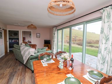 A dining table set with plates glasses and a wine bottle next to a green sofa and sliding glass doors showing an outdoor grassy area at Dol Blodau in Newbridge-On-Wye
