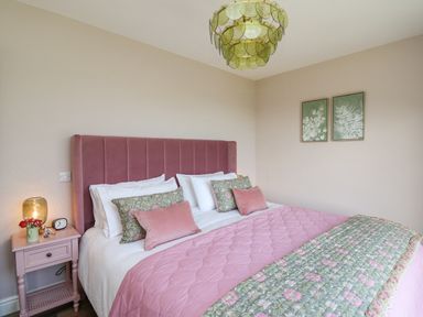 A bedroom with a pink padded headboard bed pink and floral bedding a pink nightstand with a lamp flowers and clock and two framed botanical prints on the wall at Dol Blodau in Newbridge-On-Wye
