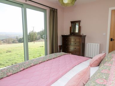 A bedroom with a pink bedspread wooden dresser with mirror and large sliding glass door overlooking a grassy area at Dol Blodau in Newbridge-On-Wye