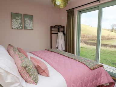 A bedroom with a bed with pink bedding two framed botanical prints on the wall a wooden standing rack with robes and a sliding glass door looking out to a grassy hill at Dol Blodau in Newbridge-On-Wye