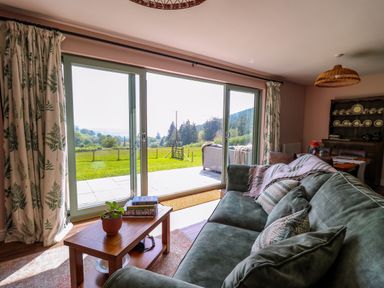 A living room with a green sofa, wooden coffee table, and large sliding glass doors showing an outdoor patio and field at Dol Blodau in Newbridge-On-Wye