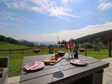 A table with red wine glasses plates bread and flowers on a patio overlooking green fields and hills at Dol Blodau in Newbridge-On-Wye