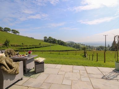 An outdoor patio with wicker furniture and table set with drinks overlooking a green field and hills at Dol Blodau in Newbridge-On-Wye