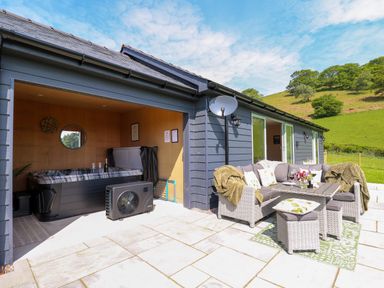 An outdoor patio with a hot tub inside a covered area and a wicker sofa set with cushions and a table on a tiled floor at Dol Blodau in Newbridge-On-Wye