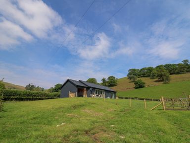 A blue cabin on a grassy hill with a fenced yard and trees in the background at Dol Blodau in Newbridge-On-Wye