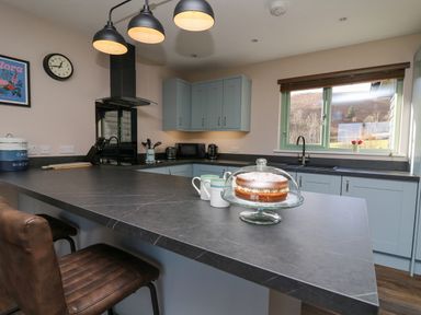 A kitchen with blue cabinets large countertop a cake on a glass stand mugs hanging lights and a window overlooking a garden at Ystrad Wen in Llysdinam near Newbridge-On-Wye