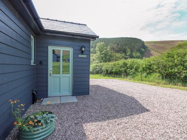 The exterior of a building with a light blue door a black wall light and a flower planter on a gravel driveway with trees and hills in the background at Ystrad Wen in Llysdinam near Newbridge-On-Wye