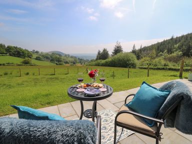 A patio with two chairs and a table set with wine glasses and food overlooking a grassy field and hills at Ystrad Wen in Llysdinam near Newbridge-On-Wye