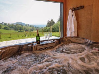 A hot tub with bubbles inside a wooden room with two white robes hanging on hooks and a bottle with two glasses on the edge overlooking a green field and hills at Ystrad Wen in Llysdinam near Newbridge-On-Wye
