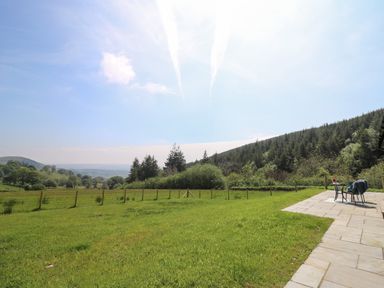 A grassy field with a fence and trees with a paved patio with table and chairs at Ystrad Wen in Llysdinam near Newbridge-On-Wye