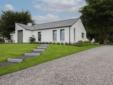 A single-story white house with a gray roof and three windows on a grassy lawn with stone steps and a gravel driveway at Shannon Vale in Woodford County Galway