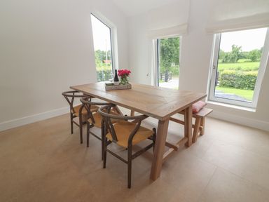 A dining area with a wooden table three chairs a bench and large windows showing a green outdoor view at Shannon Vale in Woodford County Galway