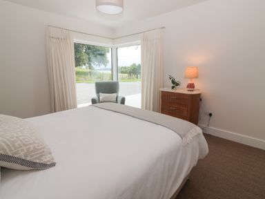 A bedroom with a bed a chair by corner windows and a wooden bedside table with a lamp at Shannon Vale in Woodford County Galway