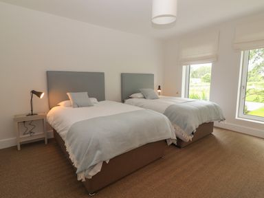 A bedroom with two beds each with a gray headboard and light blue blanket two windows and bedside tables with lamps at Shannon Vale in Woodford County Galway
