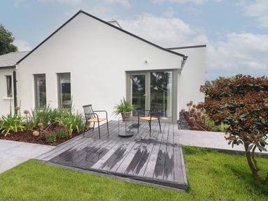 A patio with two chairs and a small table on a wooden deck outside a white house with garden plants and grass at Shannon Vale in Woodford County Galway