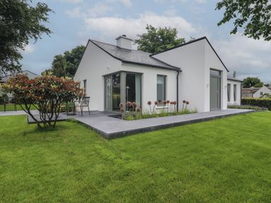 A white single-story house with large windows and a tiled patio surrounded by green grass and small bushes at Shannon Vale in Woodford County Galway