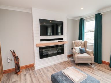 A living room with a mounted TV above a fireplace an armchair with cushions green curtains and a tray on an ottoman at 2 Apple Tree in Beadnell