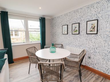 A dining area with a round white table and four wicker chairs next to a window with teal curtains and patterned wallpaper at 2 Apple Tree in Beadnell