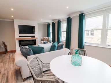 A living room with a sofa, armchair, round table with a green glass vase, and windows with teal curtains at 2 Apple Tree in Beadnell