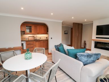 An open living and dining area with a round dining table and chairs a sofa with cushions a wall-mounted TV and a kitchen visible through an archway at 2 Apple Tree in Beadnell