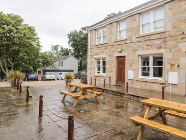 An outdoor patio area with wooden picnic tables in front of a stone building with a wooden door and windows at 2 Apple Tree in Beadnell
