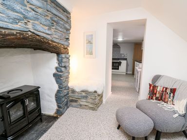 A living room with a stone fireplace a grey chair with an ottoman and a view into a kitchen at Lane Cottage in Cardigan