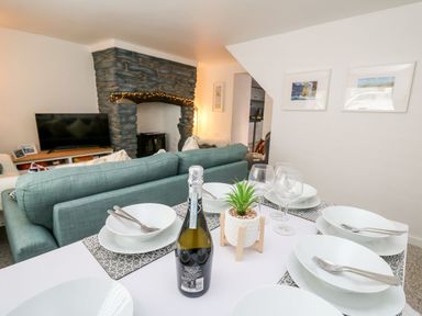 A dining table set with bowls, forks, wine glasses, a small plant, and a bottle of prosecco with a view of a living room featuring a blue sofa, stone fireplace, and a TV at Lane Cottage in Cardigan
