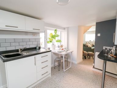 A kitchen with white cabinets a small dining table with stools and a living room visible through an open doorway at Lane Cottage in Cardigan