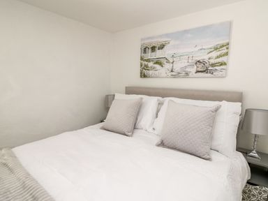 A bedroom with a white bed with two gray pillows a painting of a beach above the bed and two bedside tables with silver lamps at Lane Cottage in Cardigan