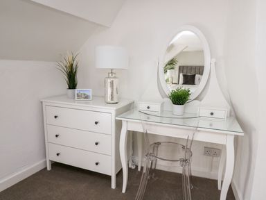 A white dressing table with a mirror and a clear chair next to a white chest of drawers with a lamp and plant at Lane Cottage in Cardigan