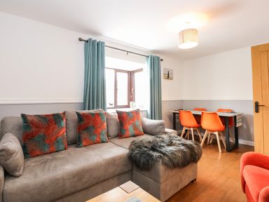 A living room with a grey sofa and orange patterned cushions near a window with blue curtains and an orange dining set at Harveys Holiday House in Hunstanton
