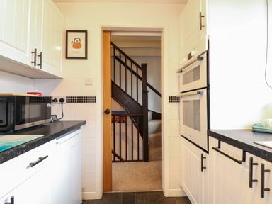A kitchen with white cabinets black handles a microwave and a double oven with a staircase visible through the doorway at Harveys Holiday House Hunstanton