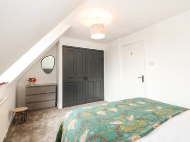 A bedroom with a bed covered with a patterned green blanket a gray chest of drawers with flowers a round mirror gray double closet doors a white door and a small wooden and metal side table at Harveys Holiday House in Hunstanton