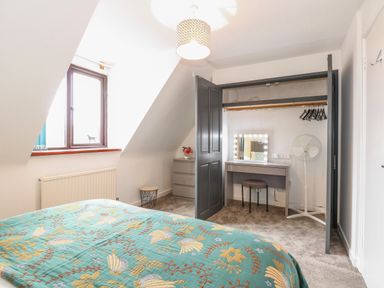 A bedroom with a bed covered in a patterned blanket a window a dresser and an open closet with a vanity table and a standing fan at Harveys Holiday House in Hunstanton