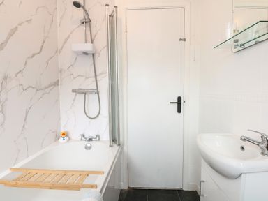 A bathroom with a bathtub with shower, a sink, a door, and a glass shelf with plastic cups at Harveys Holiday House in Hunstanton