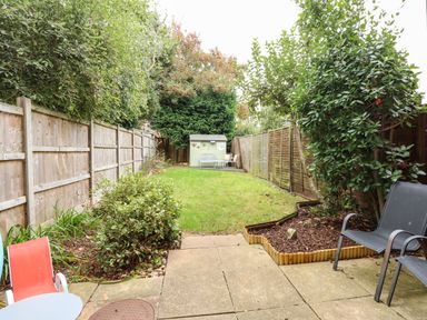 A backyard with a small green shed at the end lawn and chairs on a paved patio at Harvey's Holiday House in Hunstanton