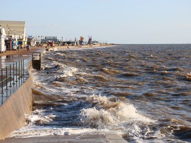 A seaside promenade with waves crashing against a sea wall and people walking near buildings at Harveys Holiday House in Hunstanton