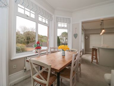 A dining room with a wooden table and six chairs next to large windows and an open kitchen area at The Anchorage in Bournemouth