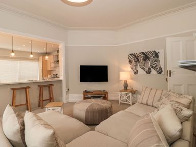 A living room with beige sofas, a round ottoman, a wall-mounted TV, and a kitchen with bar stools at The Anchorage in Bournemouth