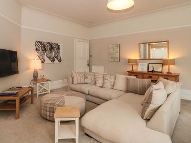 A living room with a beige sectional sofa a round ottoman two side tables a wooden console with lamps a mirror and zebra artwork on the wall at The Anchorage in Bournemouth