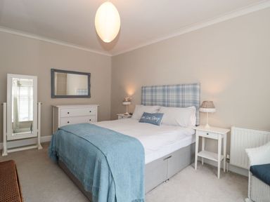 A bedroom with a bed blue throw and checked headboard two bedside tables with lamps a chest of drawers a full length mirror and a wicker chair at The Anchorage in Bournemouth